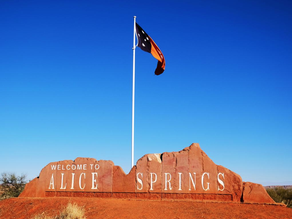Welcome to Alice Springs sign with flag on a pole against blue sky.