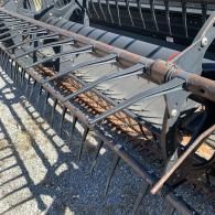 A close-up of a black combine harvester header reel with metal tines and support arms on an outdoor gravel surface.