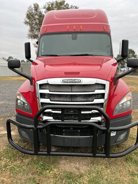 A front view of a red Freightliner semi-truck featuring a large black brush guard, parked on a dirt lot.