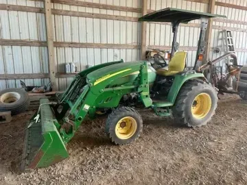 A green John Deere tractor with a front loader and yellow seat is parked inside a wooden barn.