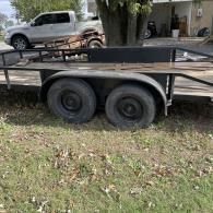 A black utility trailer with tandem wheels sits on a grassy lawn next to a white pickup truck.