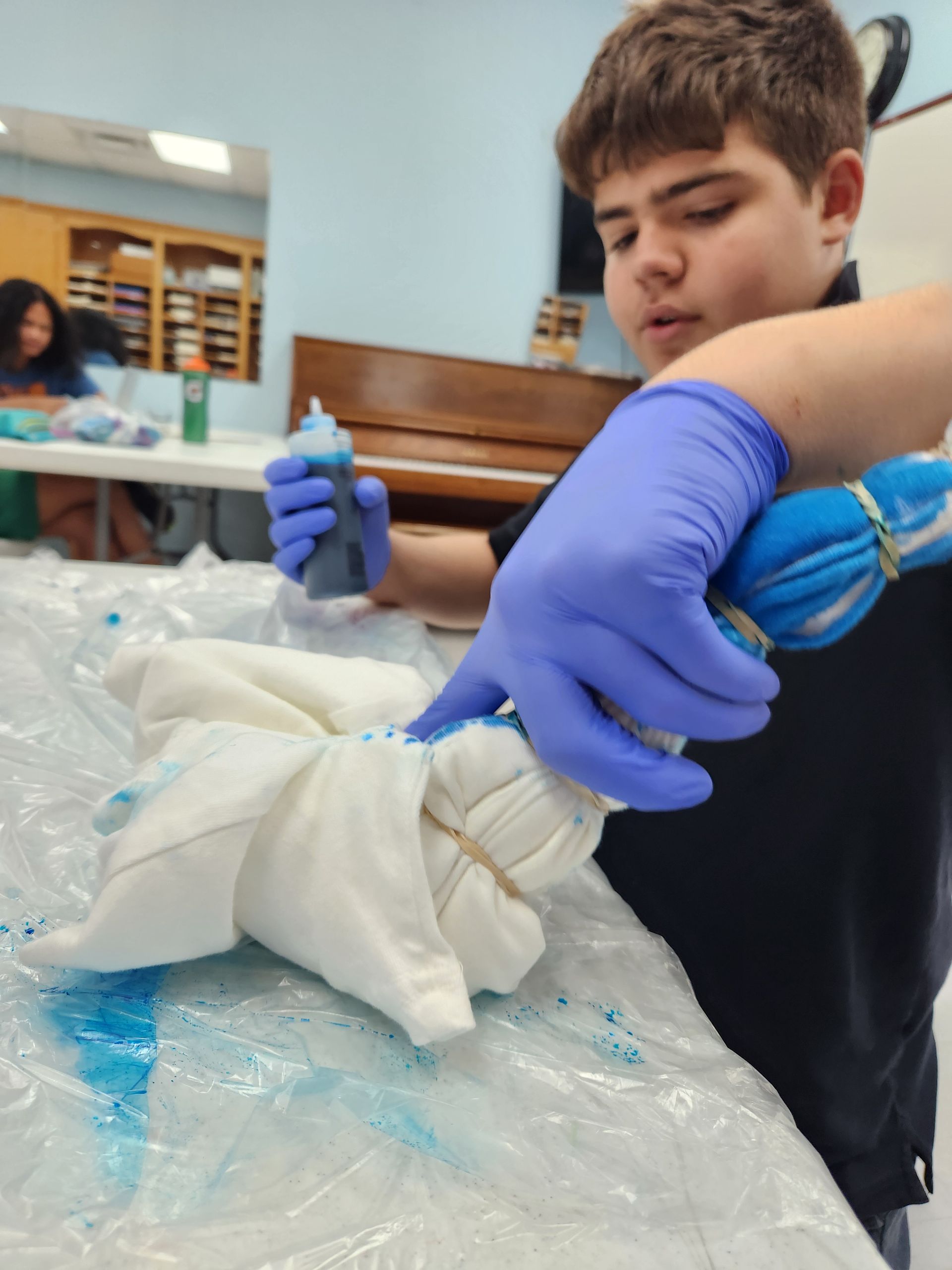 A person wearing blue gloves applies blue dye from a squeeze bottle to a tied fabric garment on a plastic-covered table.