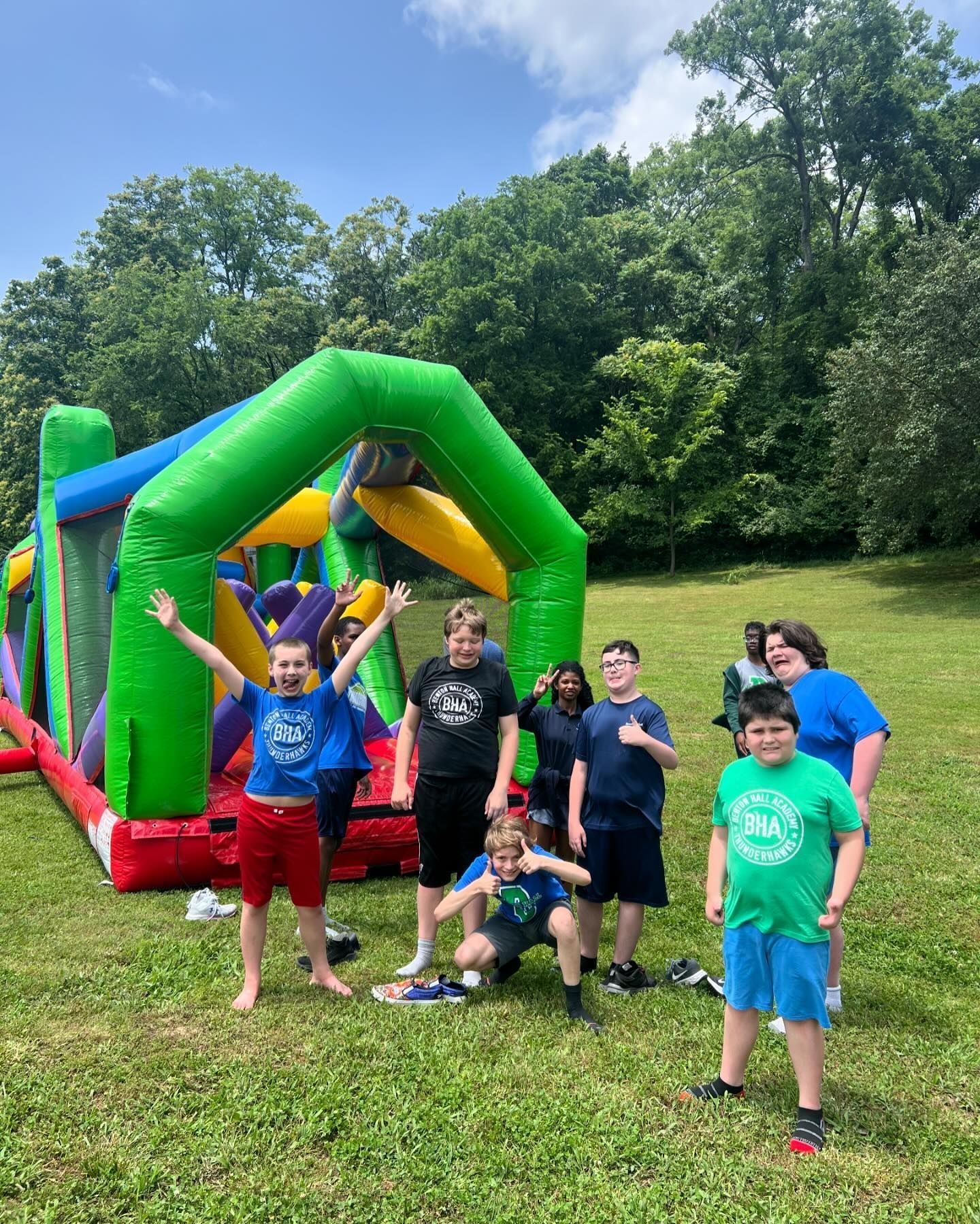 A group of young people smiling and posing in front of a colorful inflatable obstacle course in a grassy, wooded park.