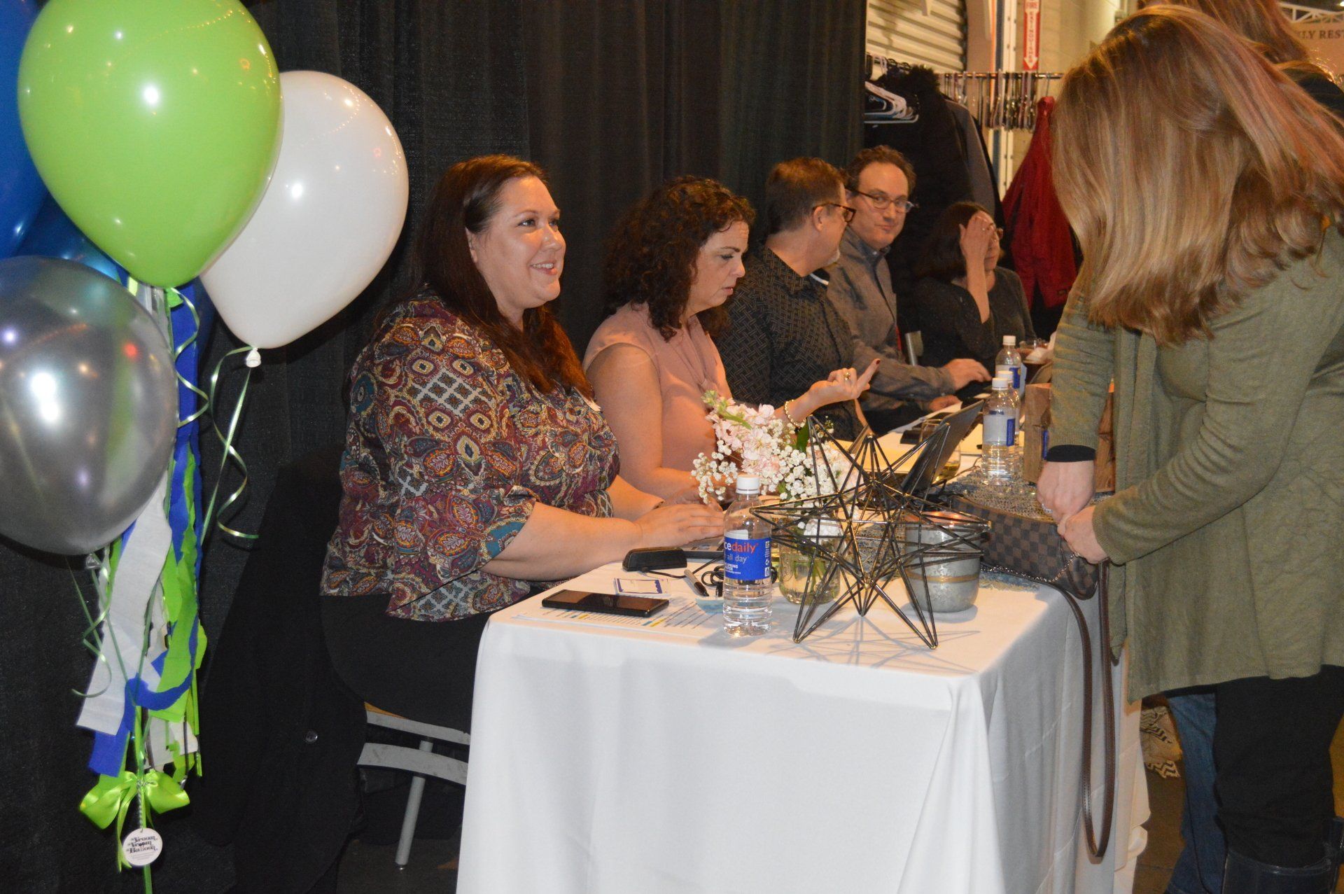 A woman smiles while signing a book for a guest at a table draped in white cloth, with others seated nearby and balloons.