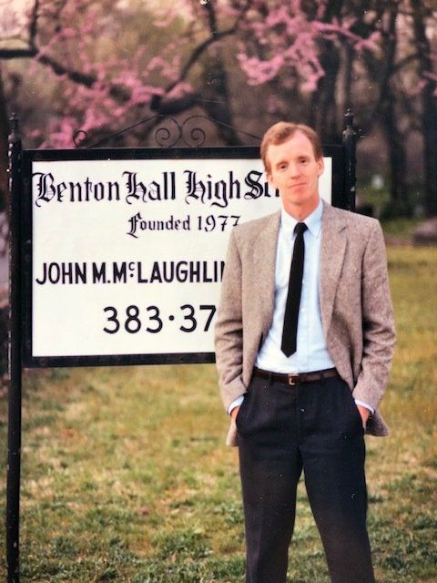 Man in blazer stands by Benton Hall High School sign.