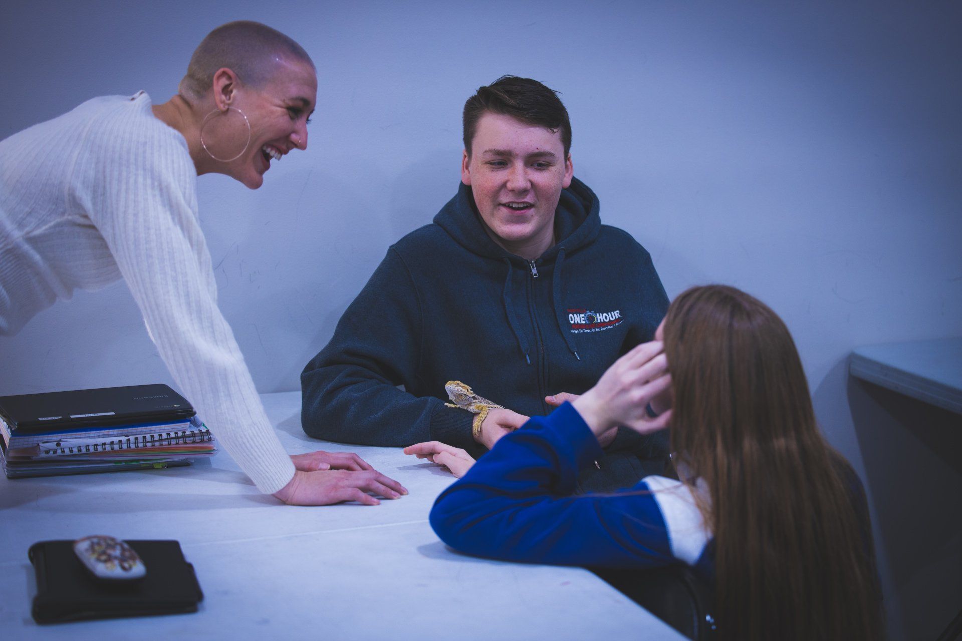 Three people gather around a white table, laughing while looking at a small reptile held in a person's hand.