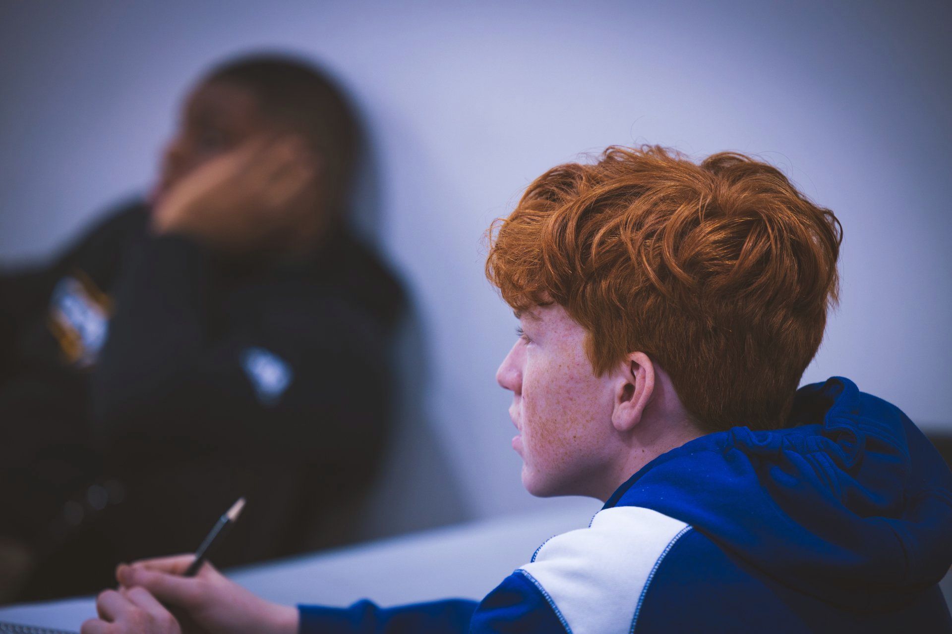 A person with red hair wearing a blue and white hoodie holds a pencil while looking off to the side in a classroom.