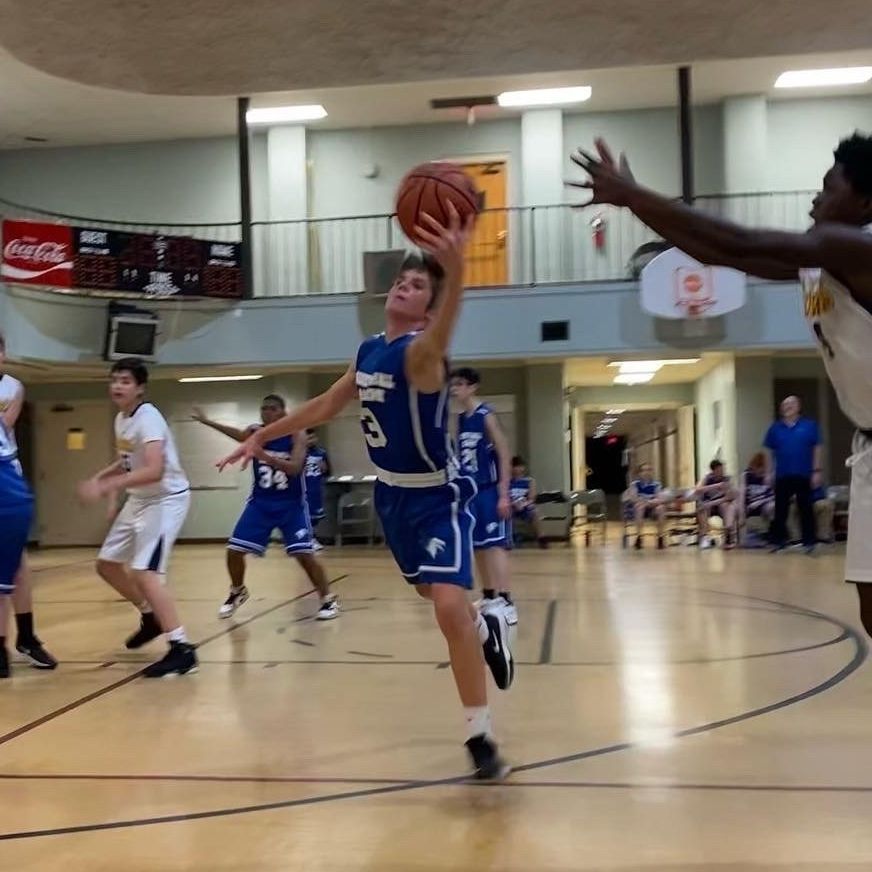 A basketball player in a blue uniform drives to the hoop for a layup while a defender reaches to block the shot.