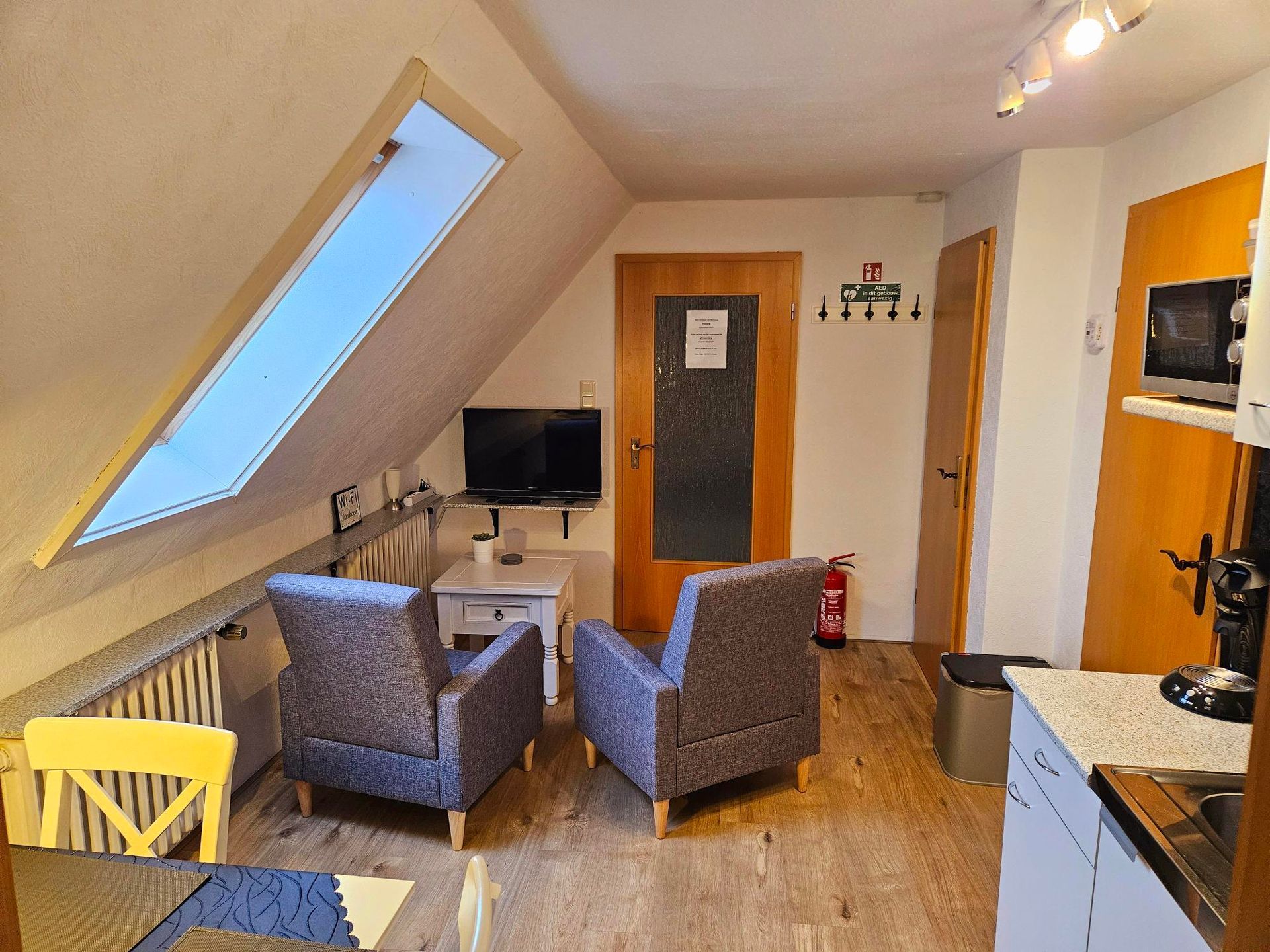 Kleine zolderwoonkamer met schuin raam, twee fauteuils, tv, bureau en keukenblad.