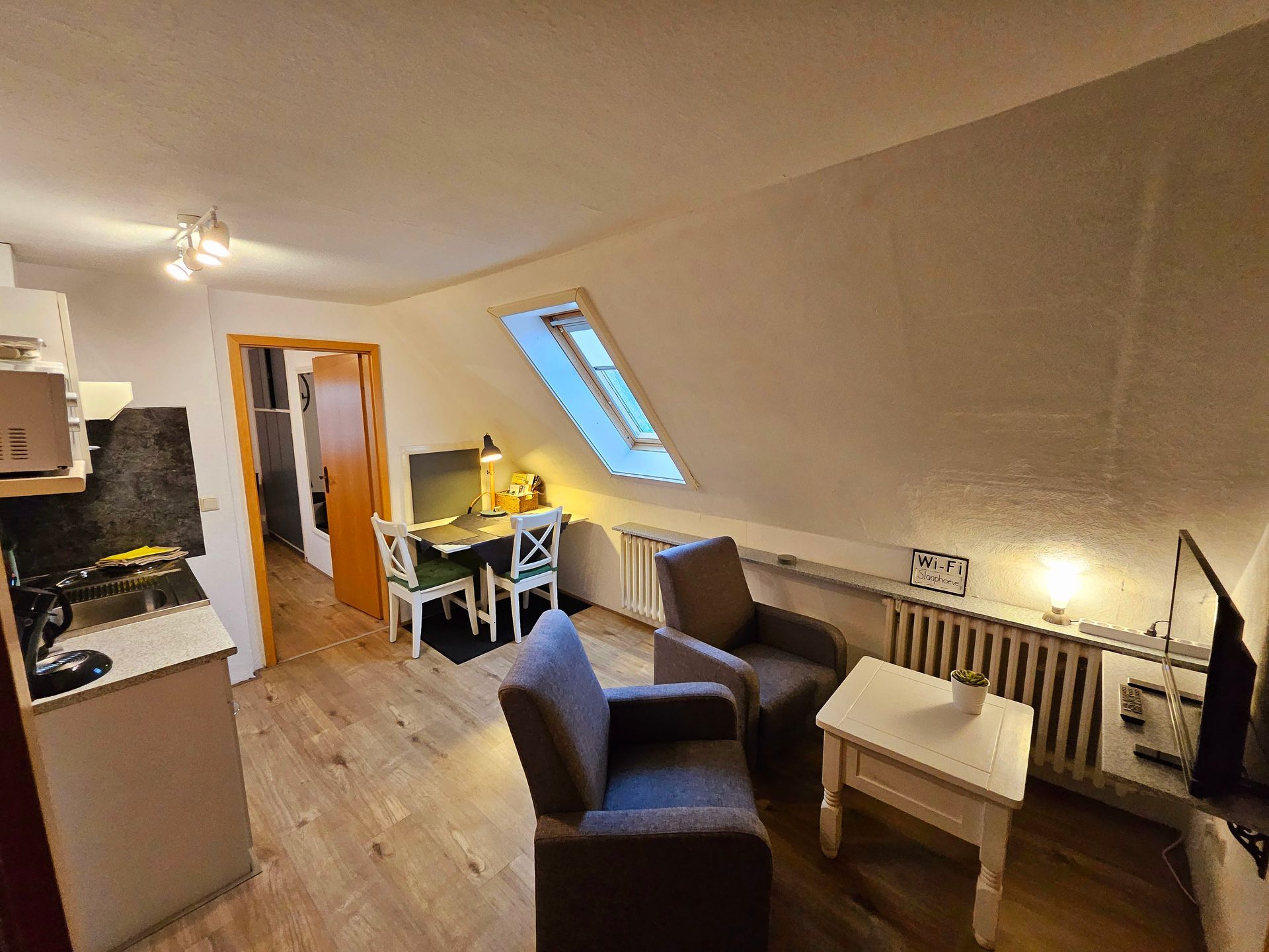 Kleine woonkamer in een appartement met twee fauteuils, een tafel, radiator, dakraam en een keukenhoek aan de achterzijde.