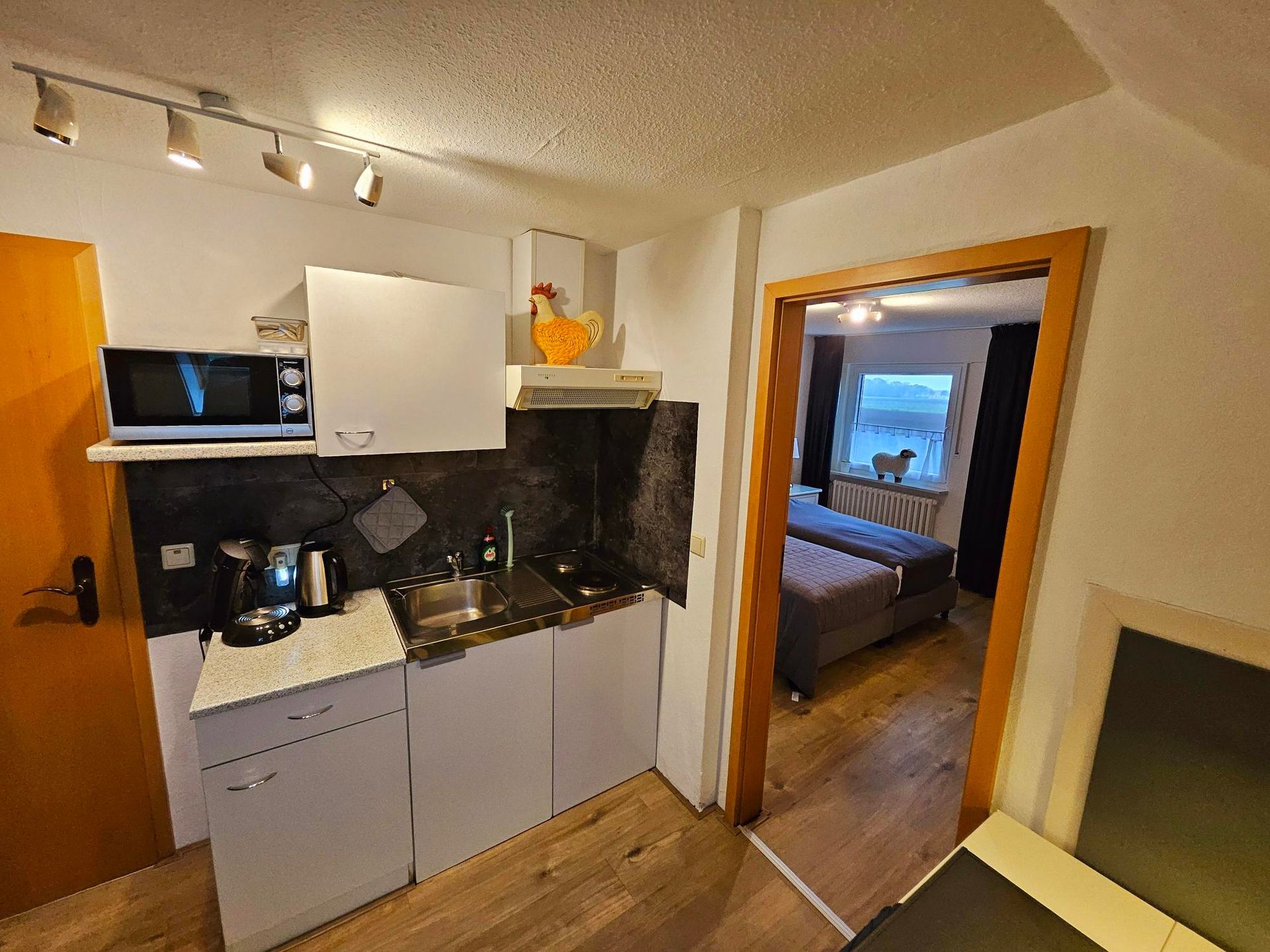 Kleine keuken met grijze kasten, zwarte aanrechtbladen en een open doorgang naar een slaapkamer.