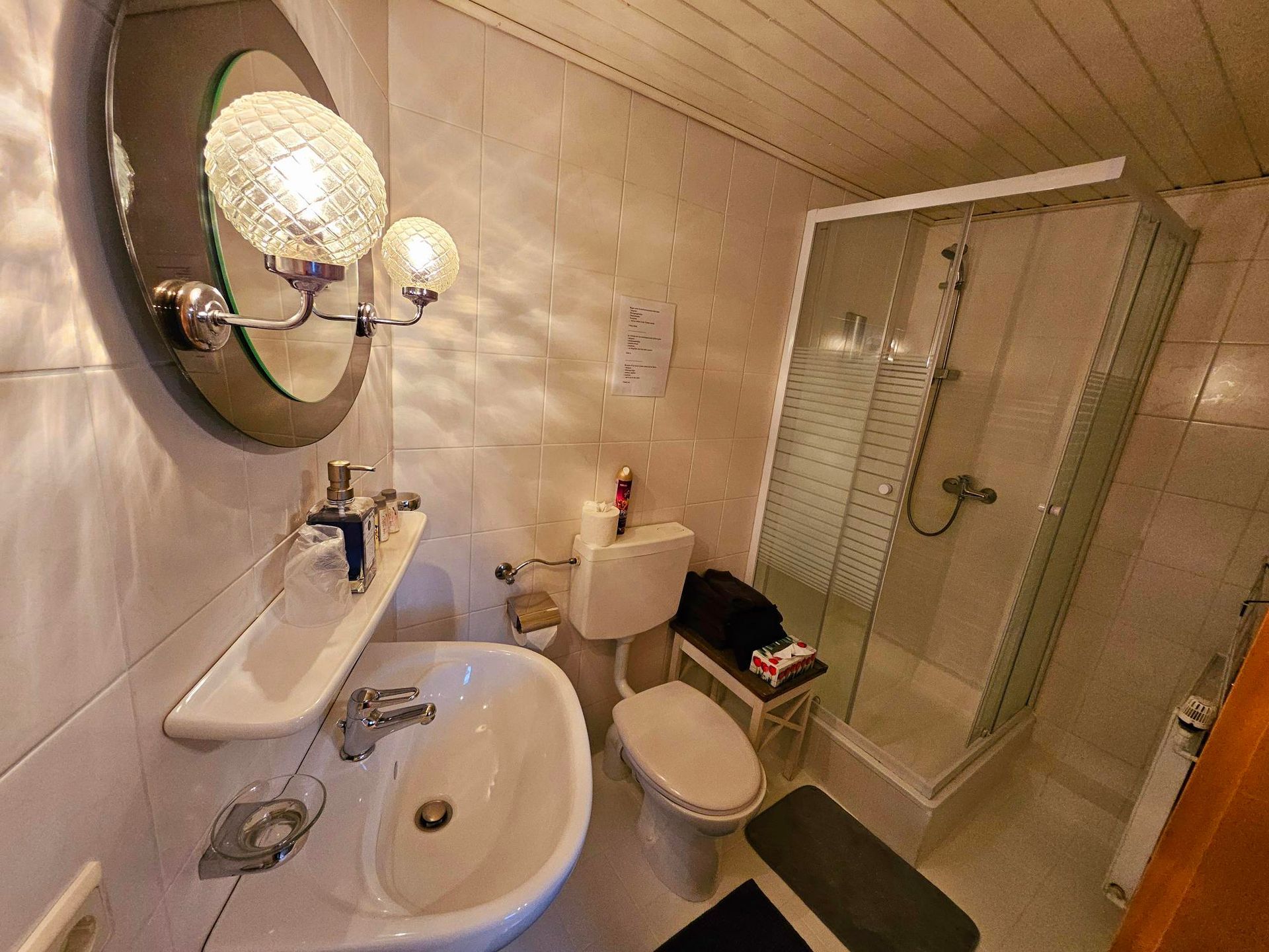 Kleine beige badkamer met wastafel, toilet, spiegel en glazen douchecabine onder warme verlichting.