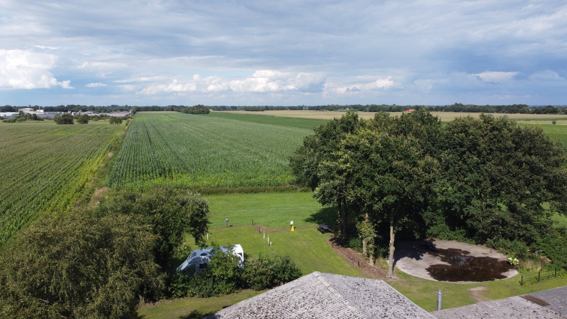 Uitgestrekt landbouwgebied onder een bewolkte hemel, met groene velden, bomen en een kleine tuin op de voorgrond.