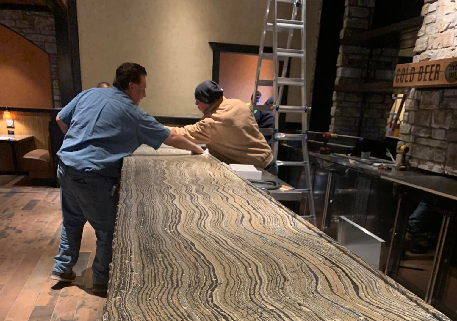 Two people installing a countertop in a bar. One on ladder, the other at counter edge.
