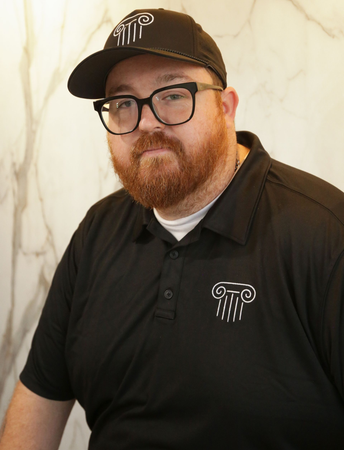 Man in black polo and cap with glasses and red beard, standing against a light marble wall.