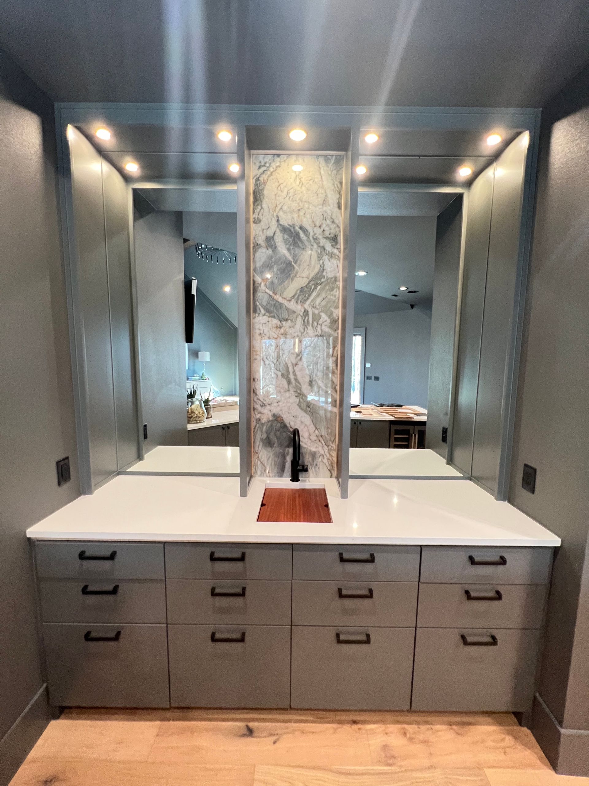 Gray built-in wet bar with mirror backsplash, white countertop, drawers, and wood sink.