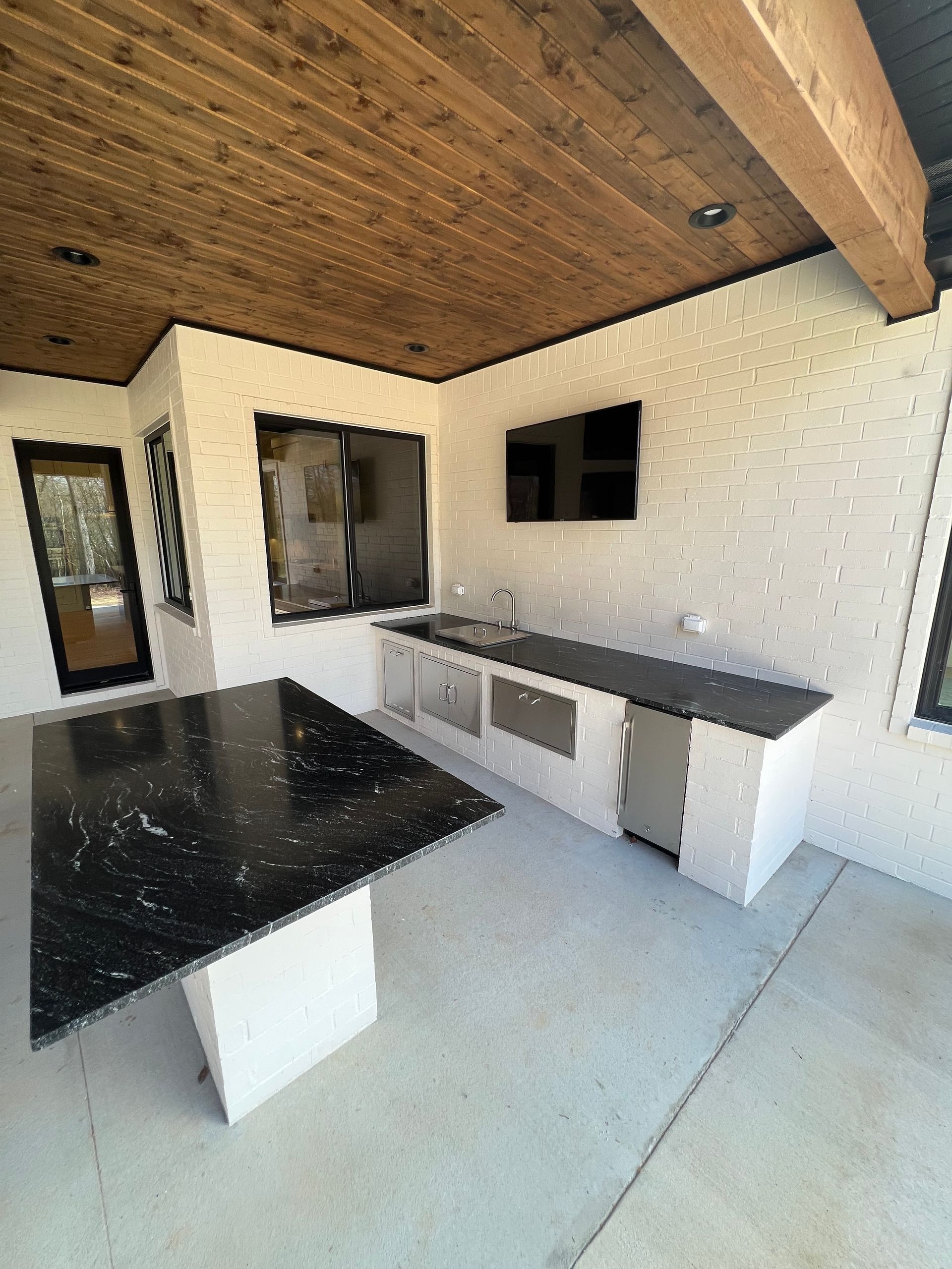 Outdoor kitchen with black countertops, a TV, and white brick walls. Wooden ceiling and concrete floor.