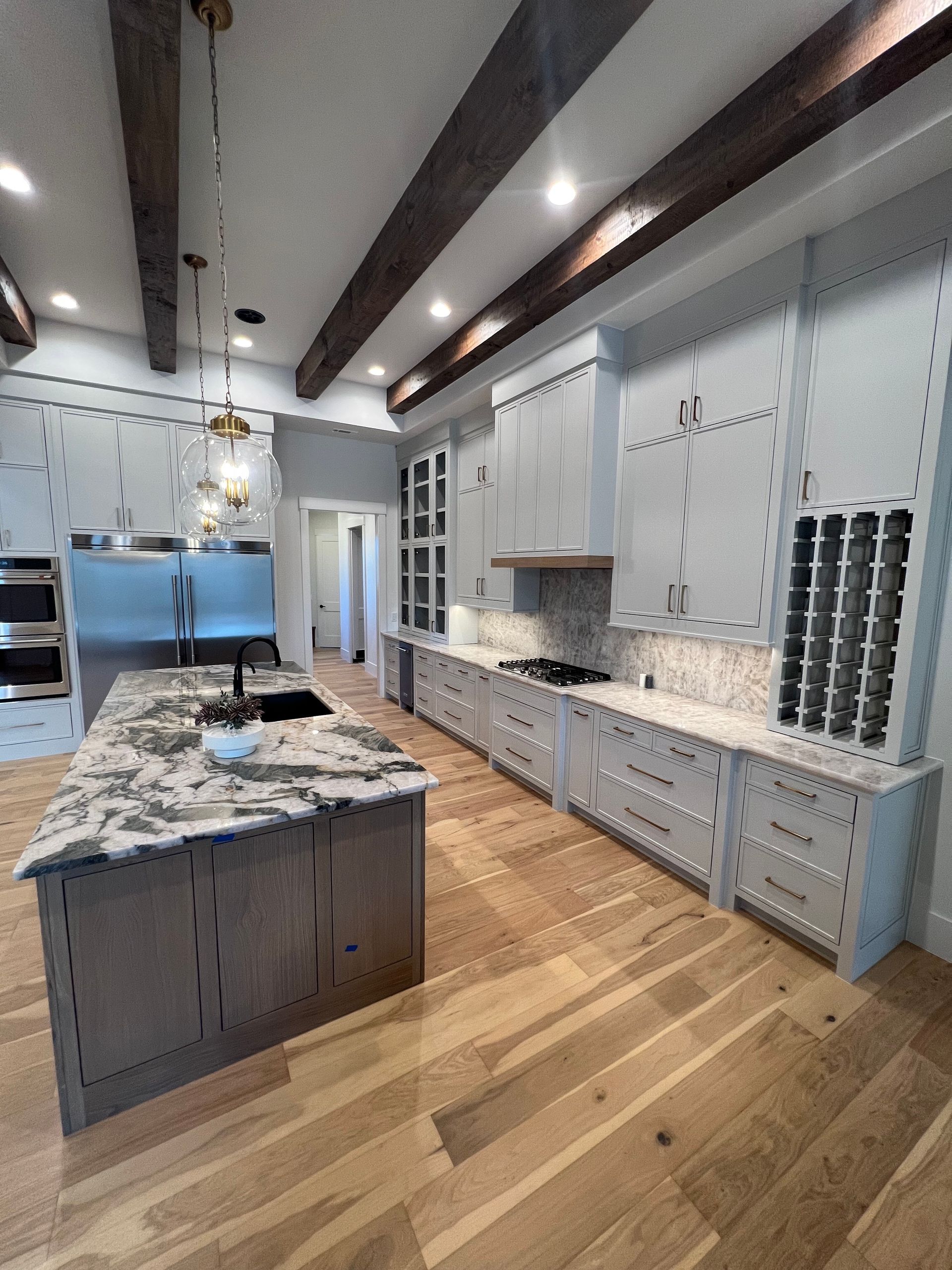 Spacious modern kitchen with light wood floors, gray cabinets, granite countertops, and wood ceiling beams.
