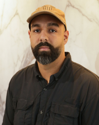 Man standing indoors against a light marble wall, wearing a tan cap and dark button-up shirt.