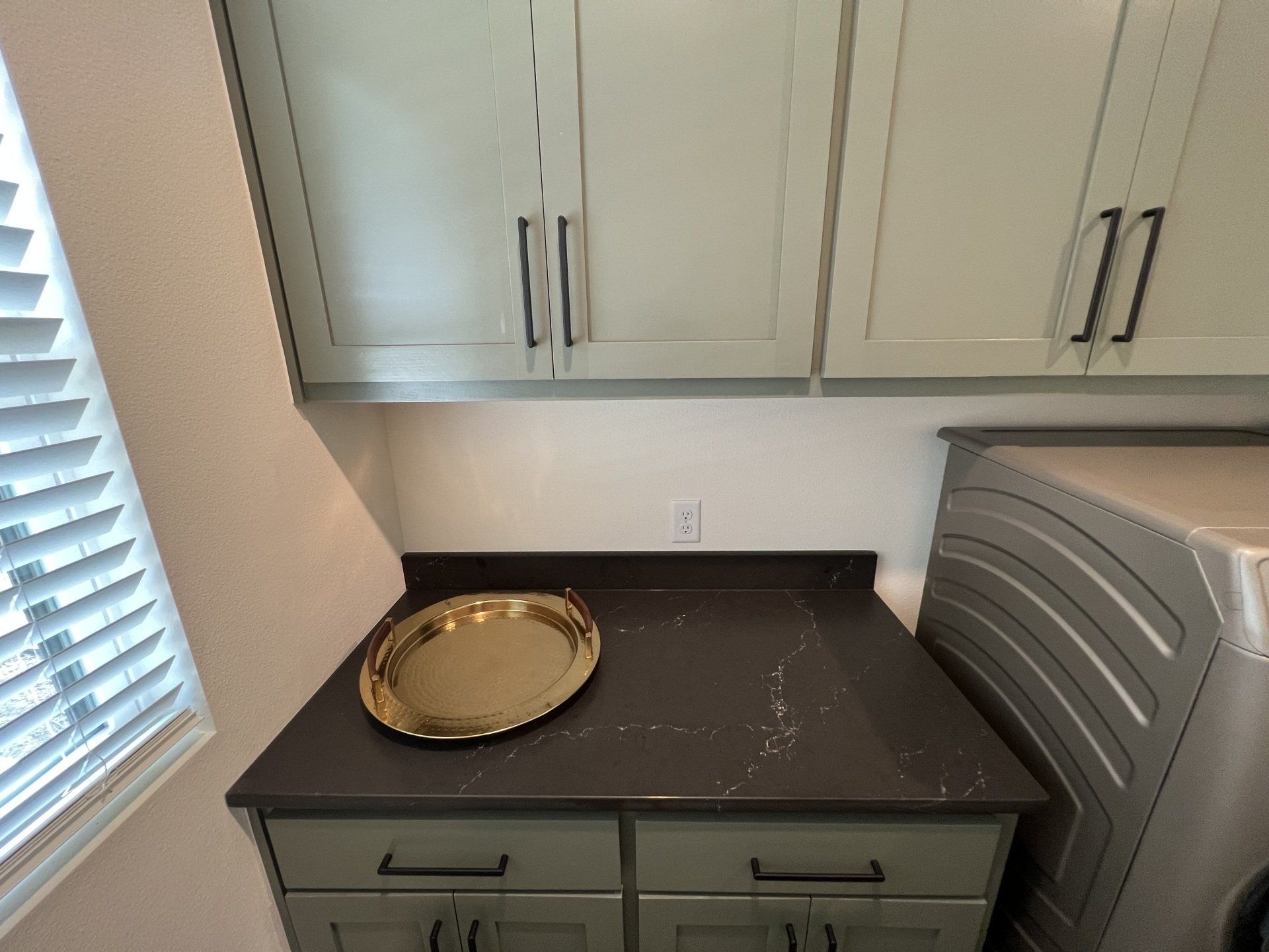 Laundry room with cabinets, counter, and a washer. Gold tray on counter, blinds to the left.