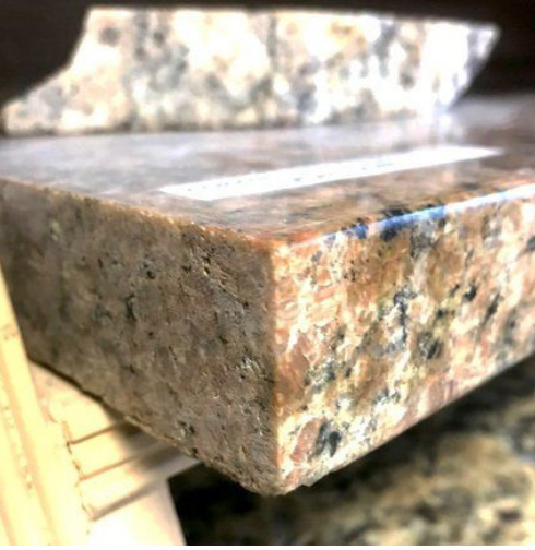 Close-up of a granite countertop sample with rough edges. Colors include tan, pink, black, and gray.