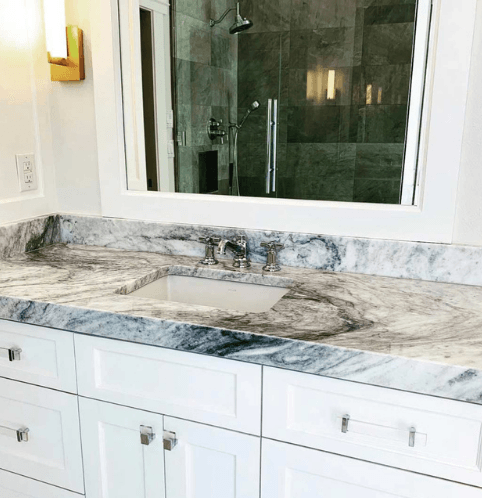 Bathroom vanity with a gray marble countertop, white cabinets, and a mirror reflecting a shower.