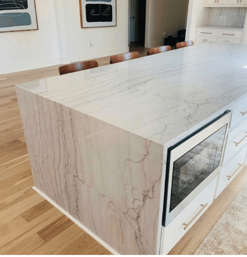 A kitchen island with a marble countertop and built-in microwave, white cabinets, and wooden floors.
