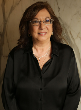Photo of a woman in a black blouse and glasses, standing against a marble wall.