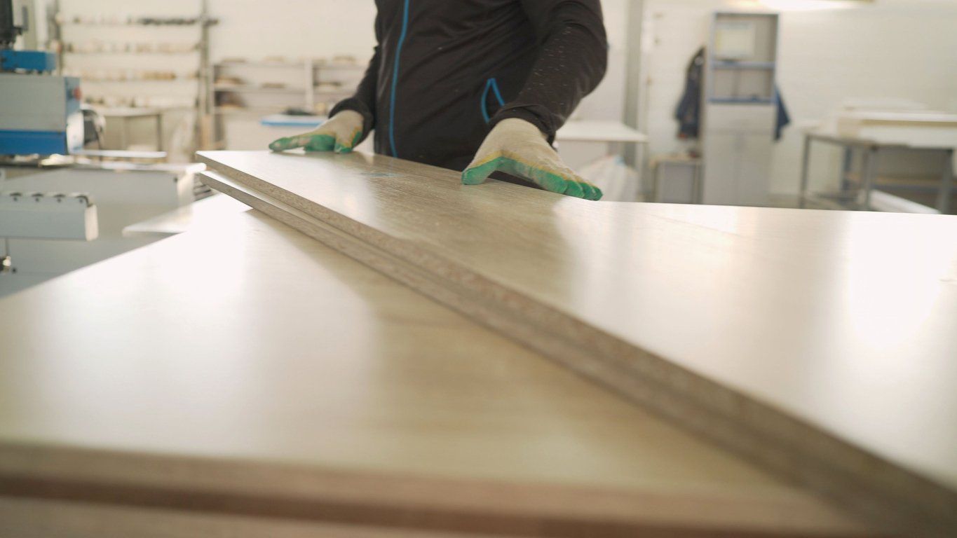 Person wearing gloves inspecting a light-colored wooden panel in a factory setting.