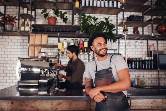 Smiling barista in apron leaning on the counter, other barista in the background, coffee shop setting.