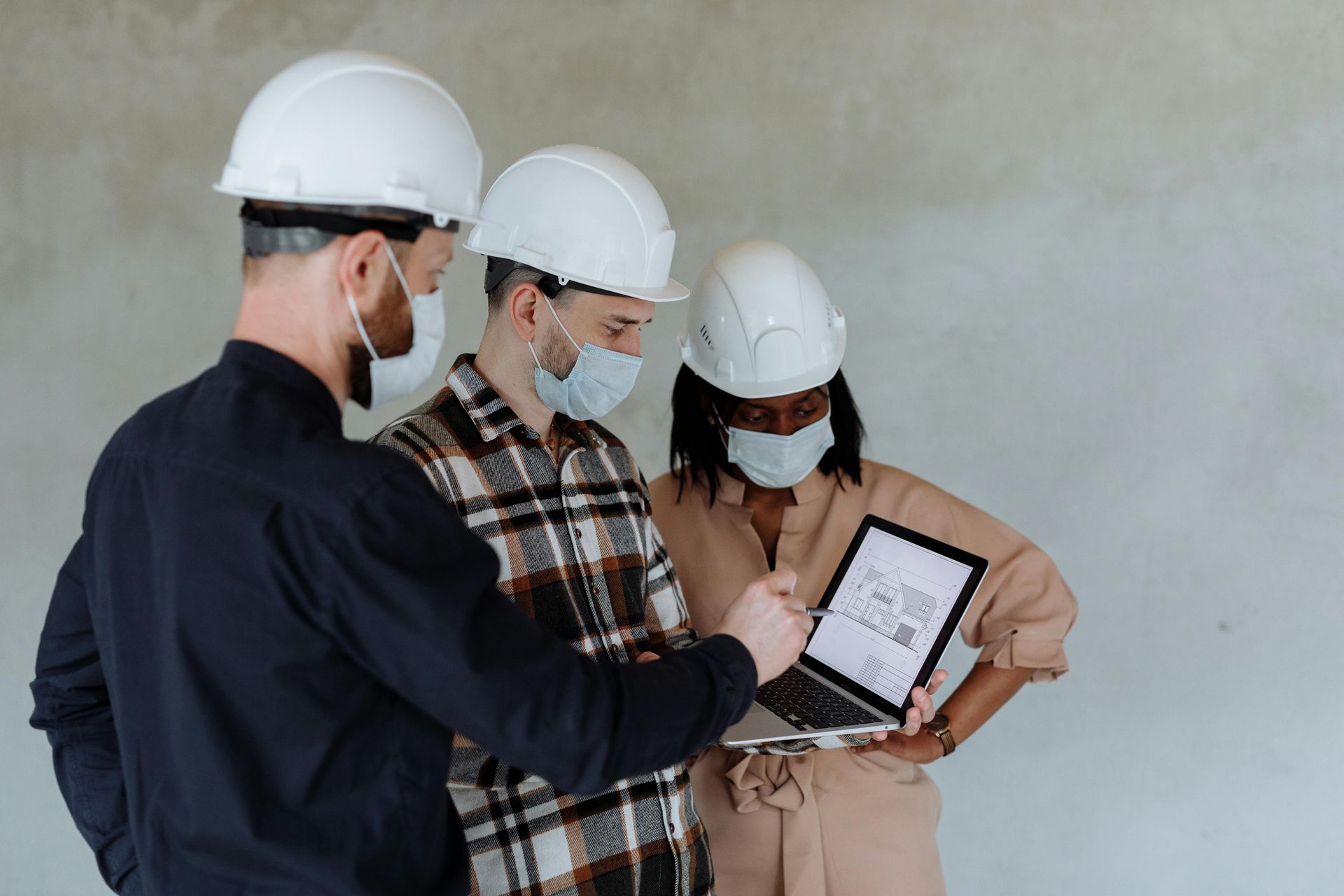 Three people in hard hats and masks looking at a tablet with a blueprint indoors.