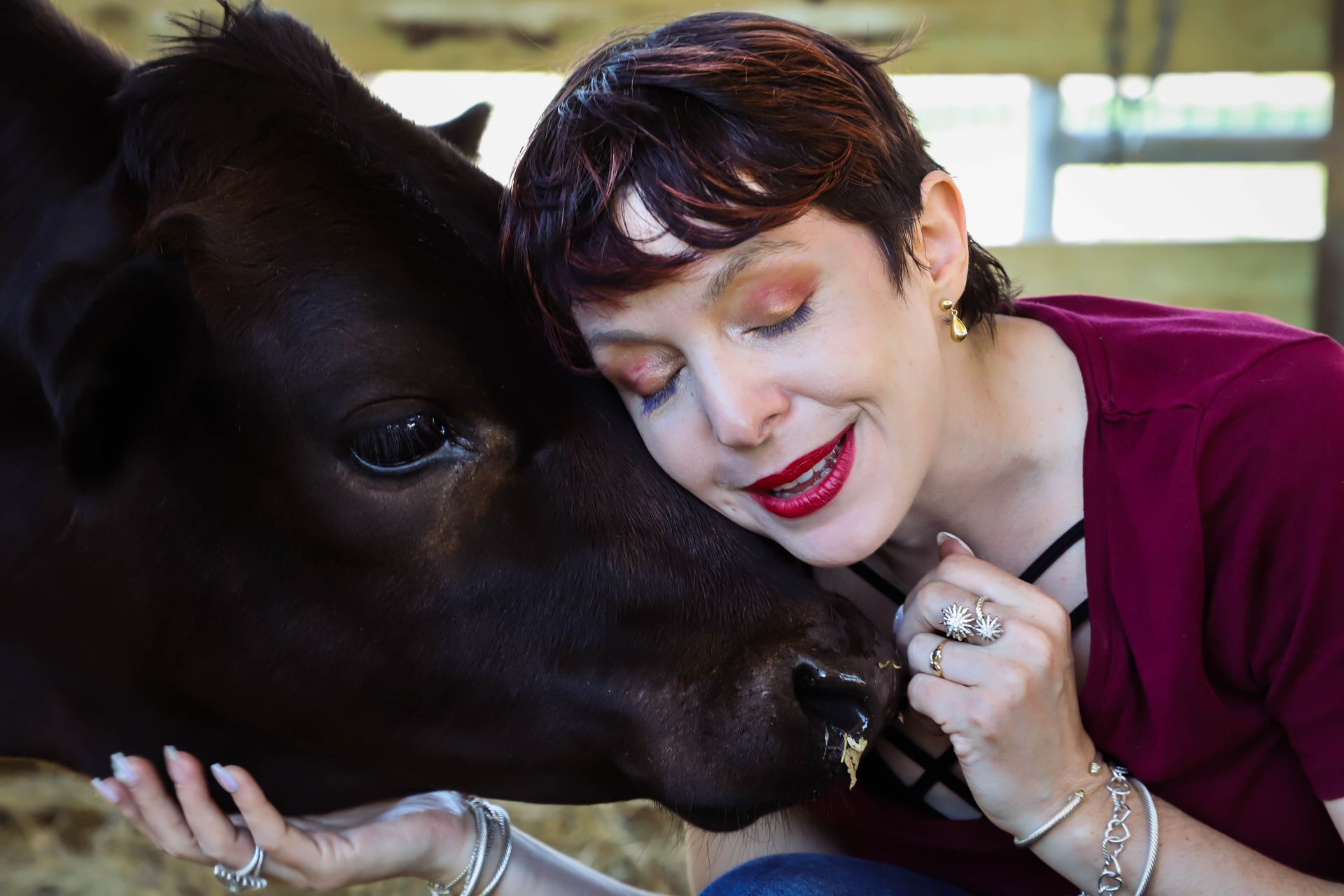Woman with short hair embraces a black cow, both with eyes closed. Indoors, barn setting.
