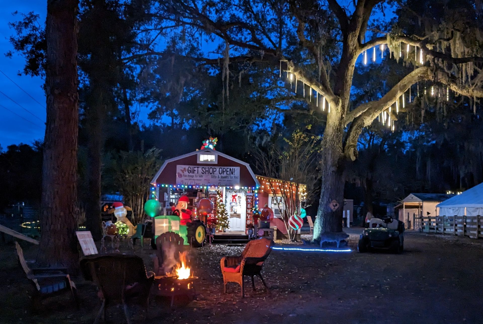 Festive barn with Christmas lights, bonfire, chairs; a tractor. Nighttime scene.