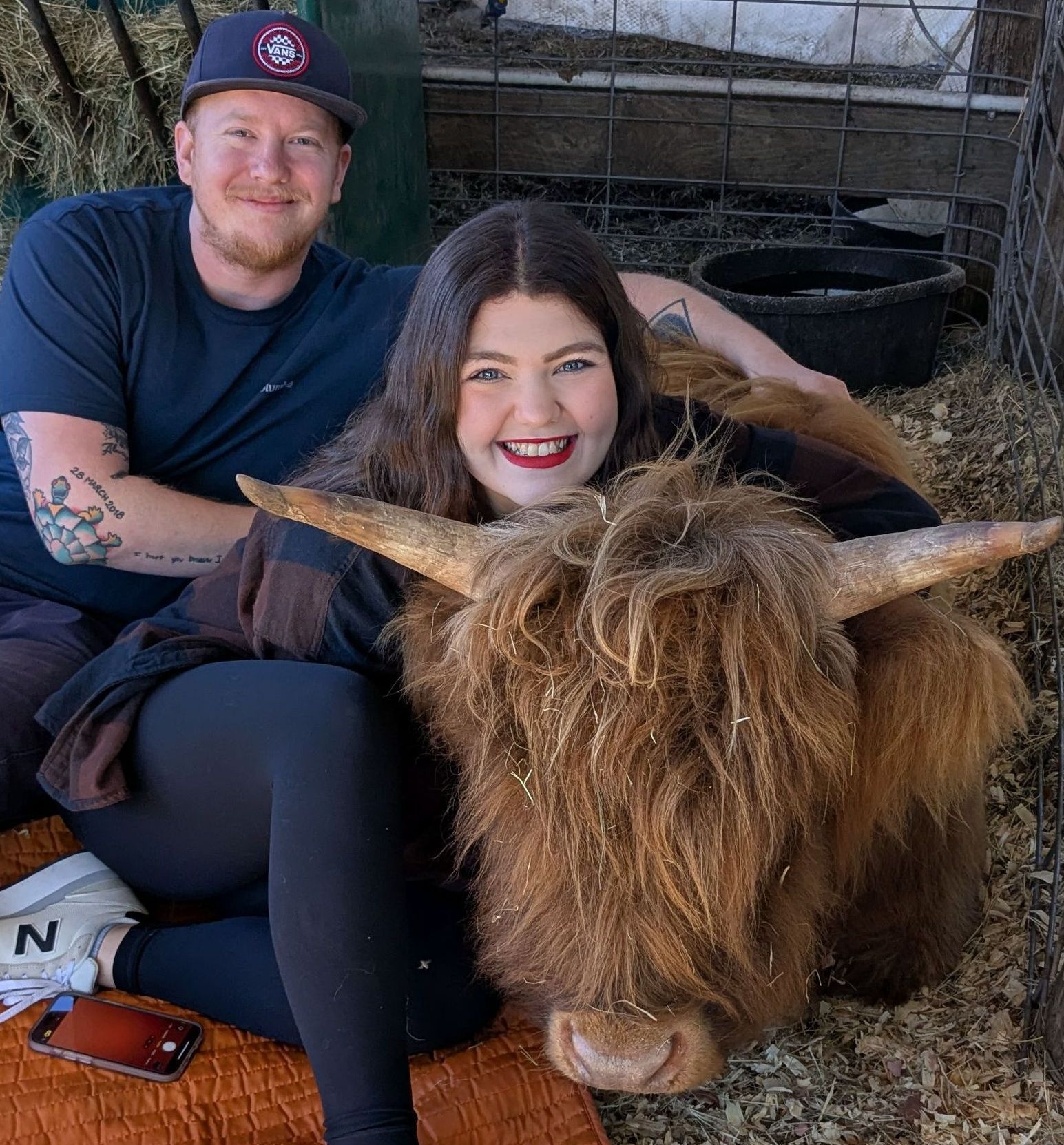 A woman and man smiling with a fluffy brown Highland cow. They are outside.