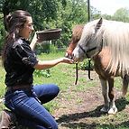 Woman kneels feeding a pony from a bowl outdoors. The pony has light blonde mane.