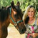 Woman with blonde and dark hair smiles next to a brown horse wearing a green halter outdoors.