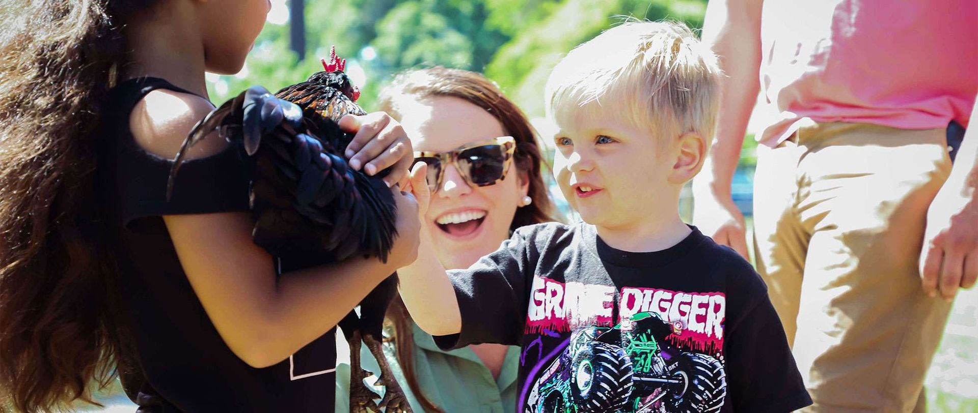 A child holds a chicken. A smiling woman and another child look at it. Outdoors in sunlight.