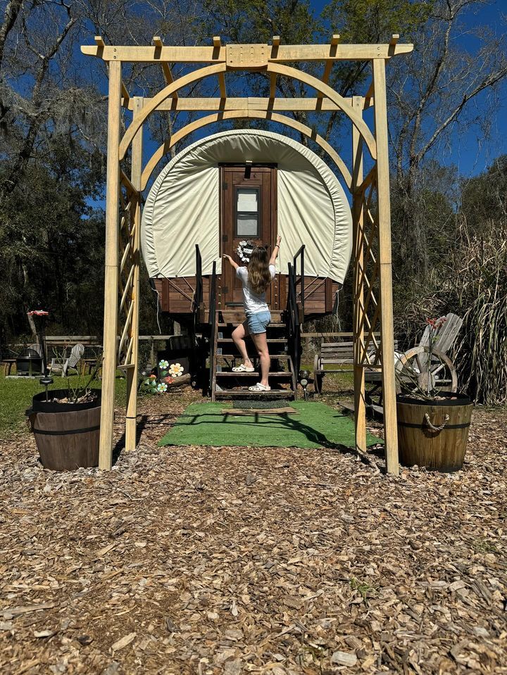A wooden arbor frames a round-roofed wagon with a person climbing the steps.