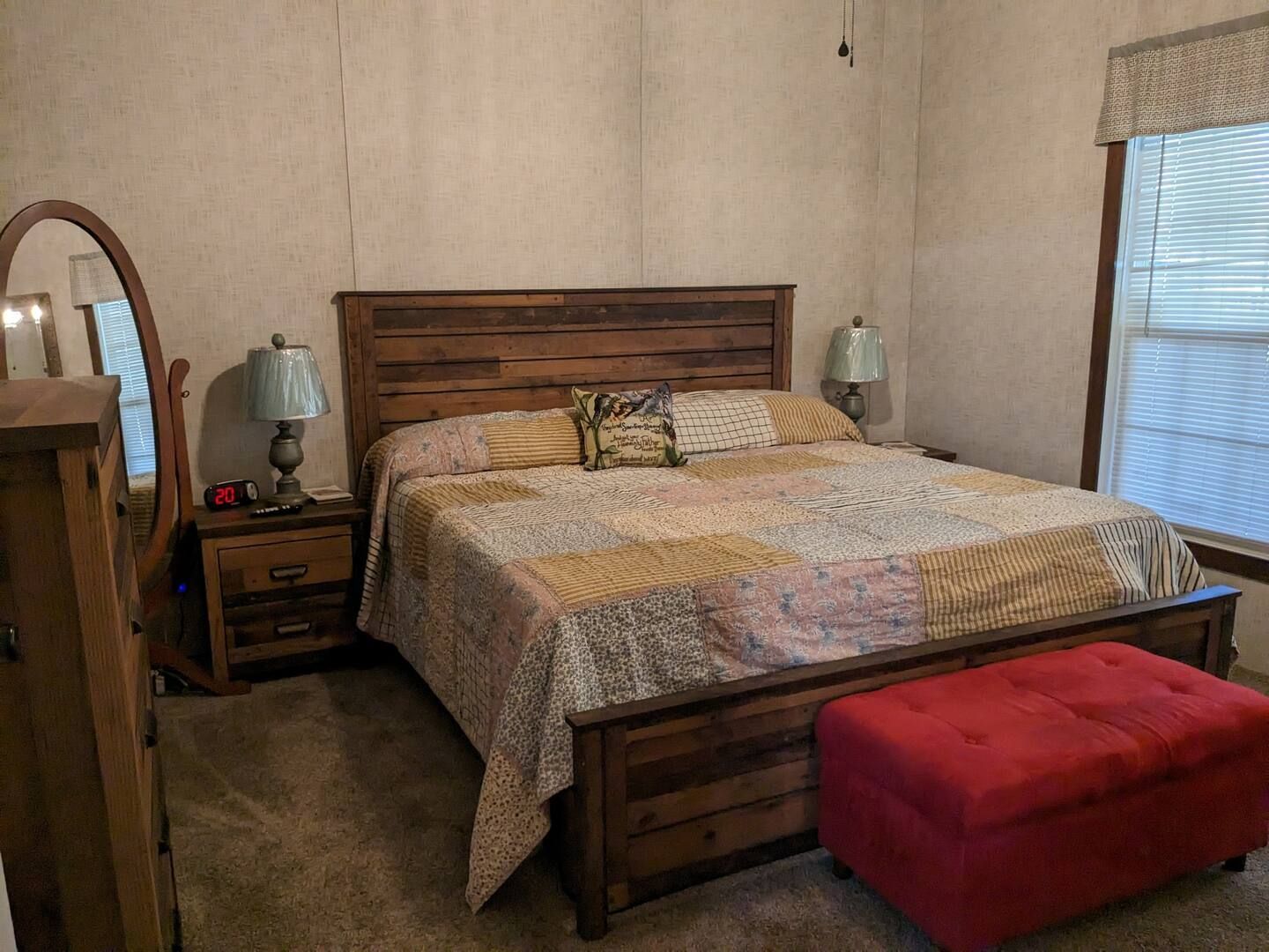Bedroom with wooden furniture and a red ottoman. A bed, nightstands, and mirror are visible.