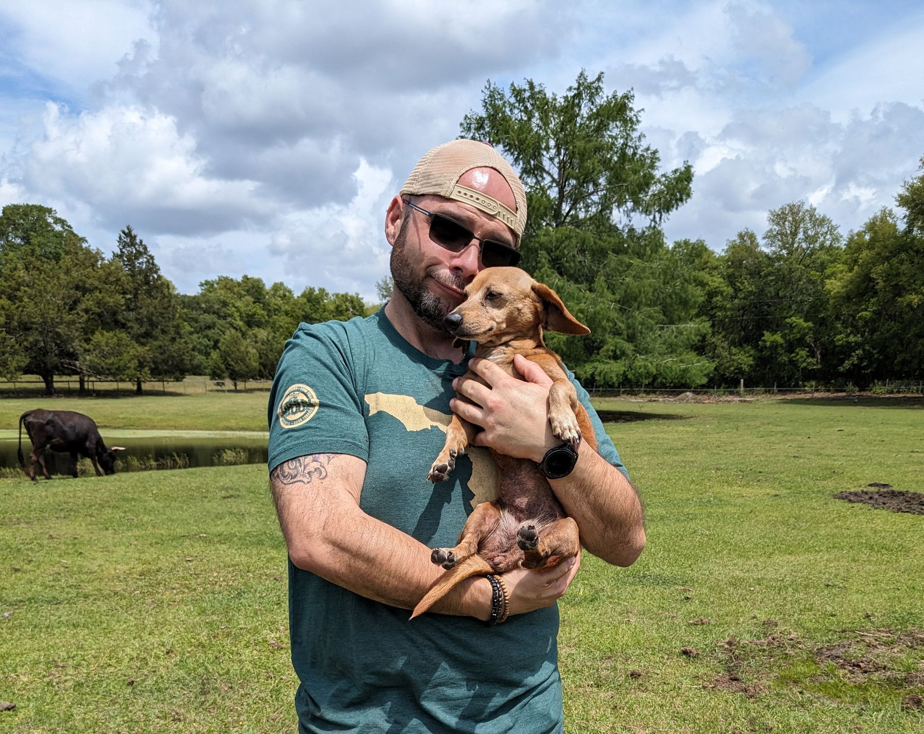 Man in cap and sunglasses holding small dog in grassy field with pond and trees.