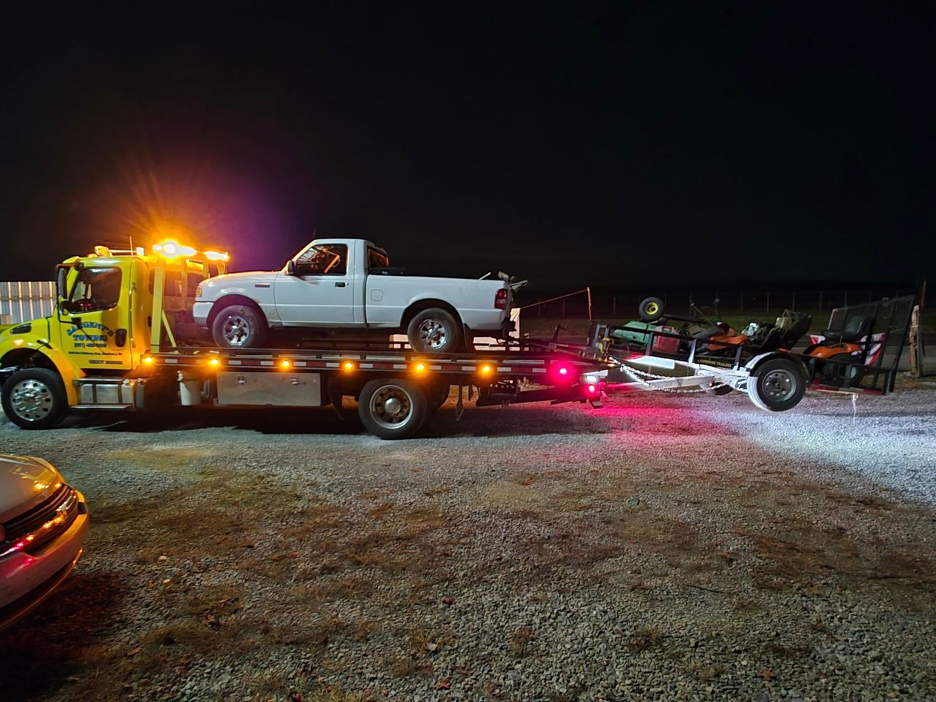 Yellow tow truck at night towing a white pickup truck and a trailer with two ATVs.