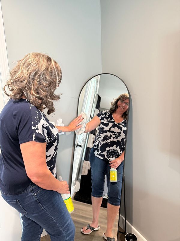 A person cleaning an arched floor mirror with a spray bottle and cloth in a room with grey walls.