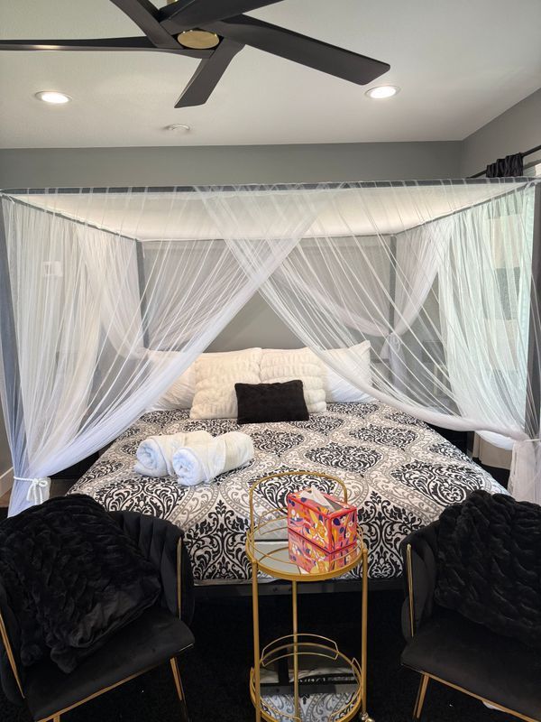 A four-poster bed with white canopy netting, patterned bedding, and two chairs in a bedroom with gray walls.