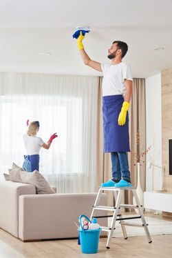 Two cleaners in white shirts and blue aprons clean a bright, modern living room; one stands on a ladder wiping the ceiling.