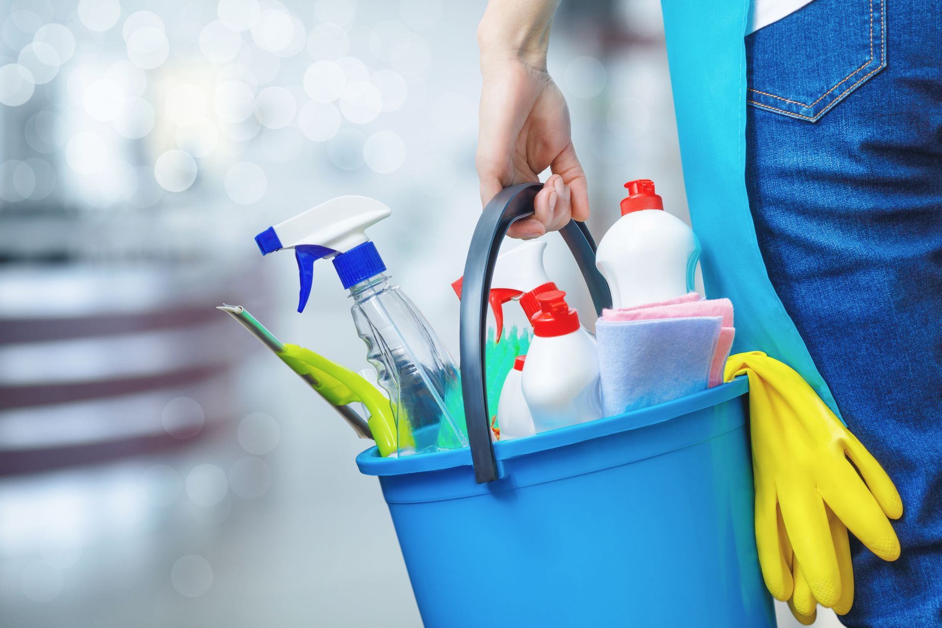 A person holding a blue bucket filled with various colorful cleaning supplies and yellow rubber gloves.