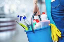 A person holding a blue bucket filled with various colorful cleaning supplies and yellow rubber gloves.