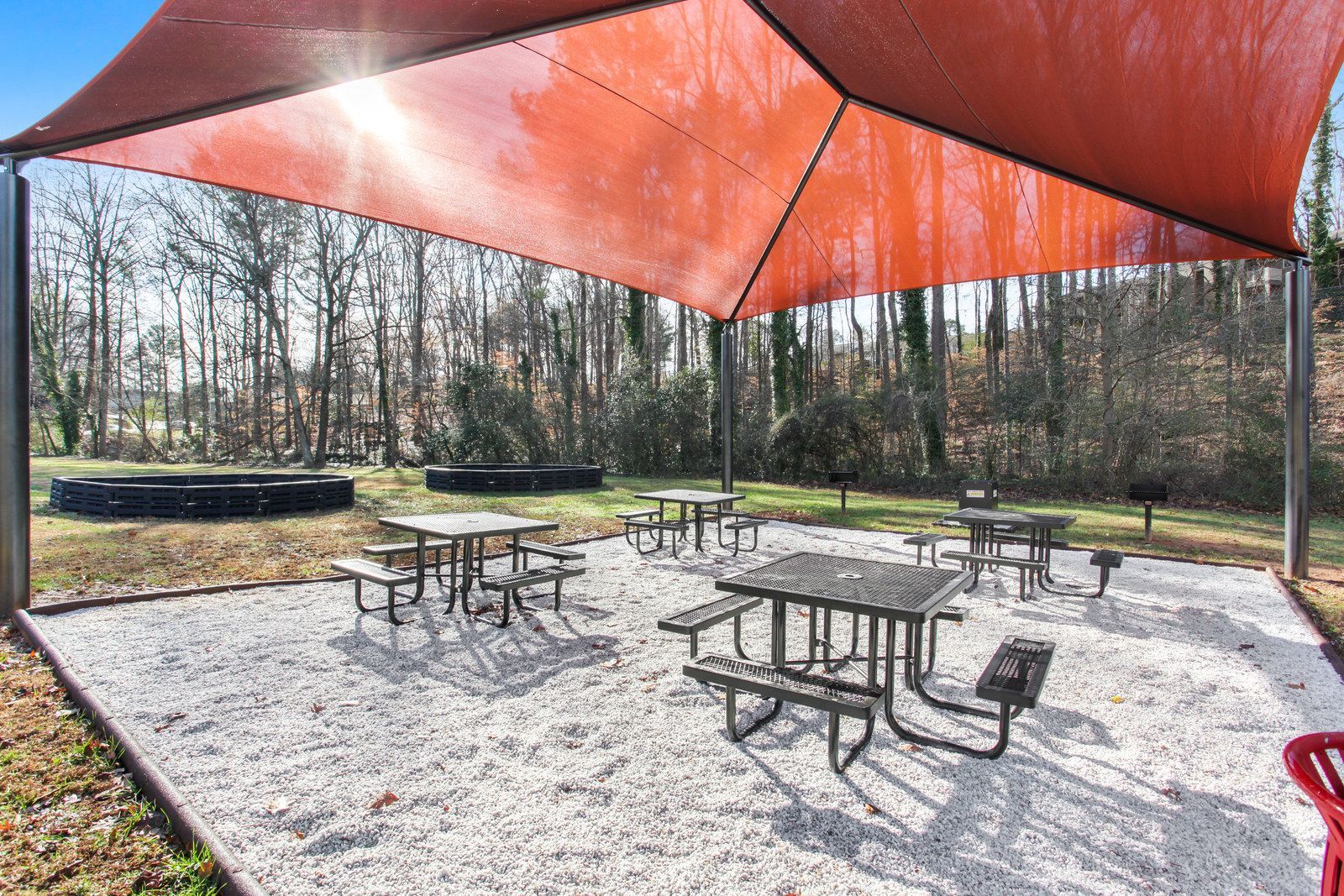 Outdoor picnic area with metal tables under a large orange shade canopy, surrounded by trees