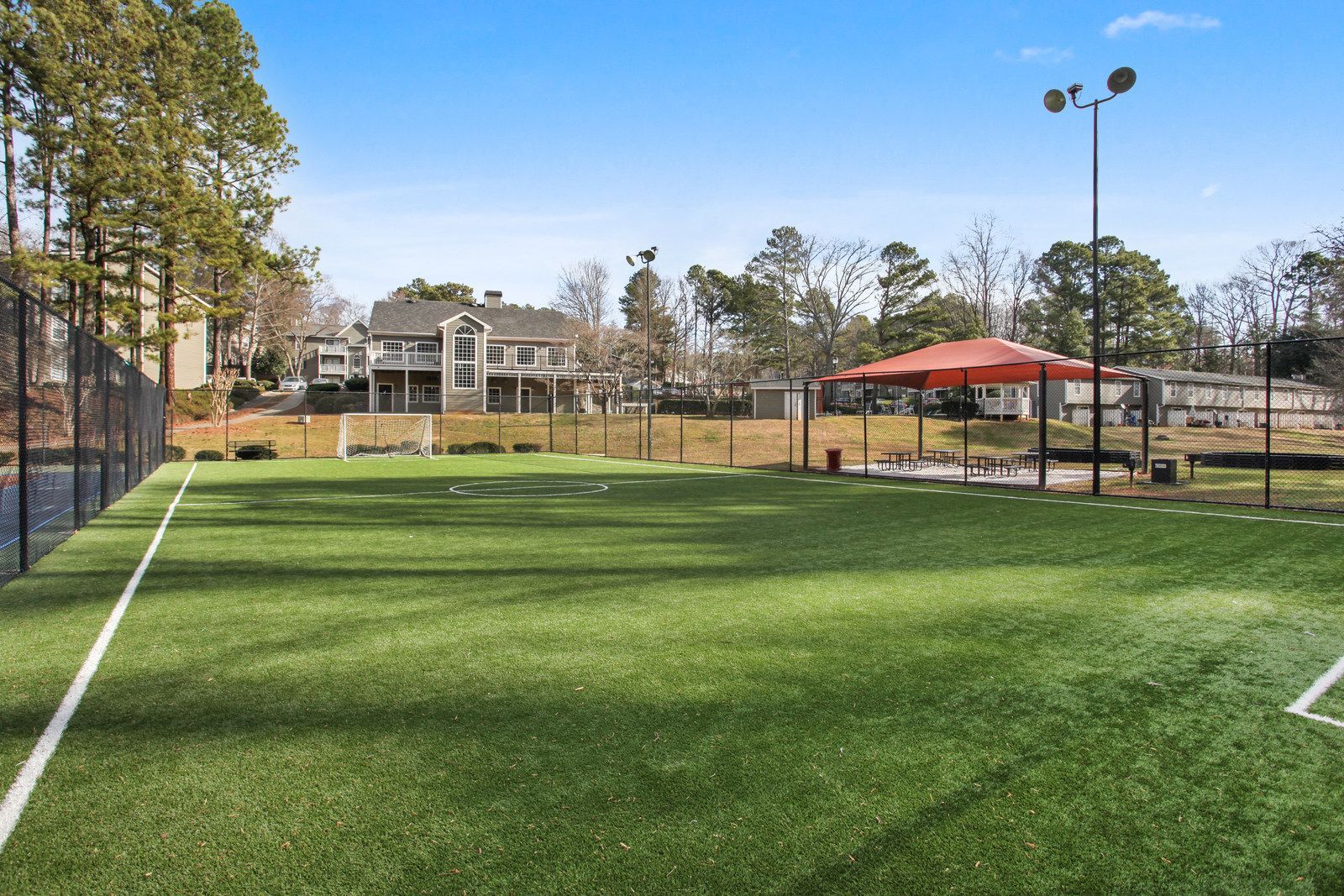 Fenced green sports field with artificial turf, red shade canopy, and apartment buildings in the background