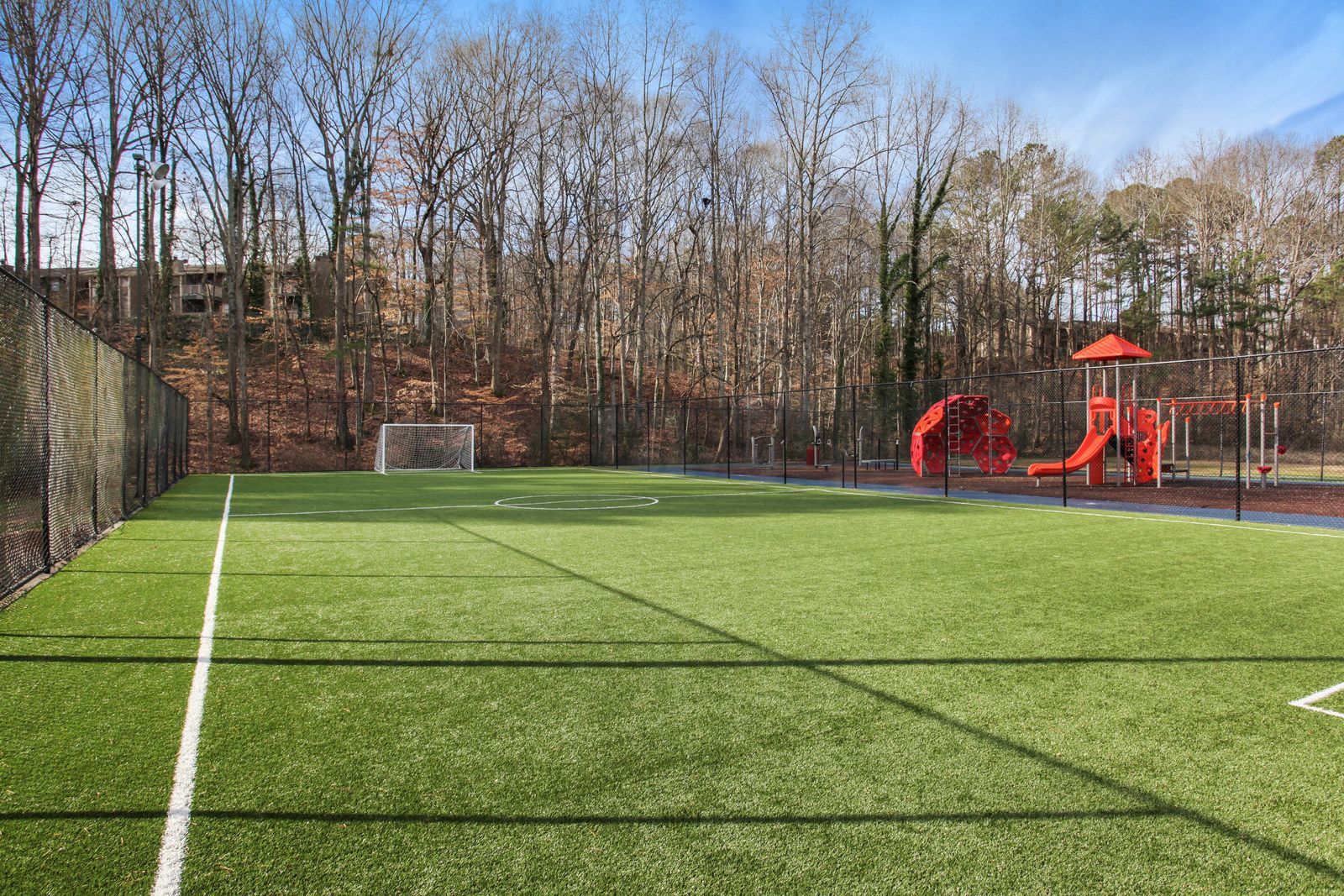 Artificial turf sports field with a soccer goal and red playground equipment beside leafless trees
