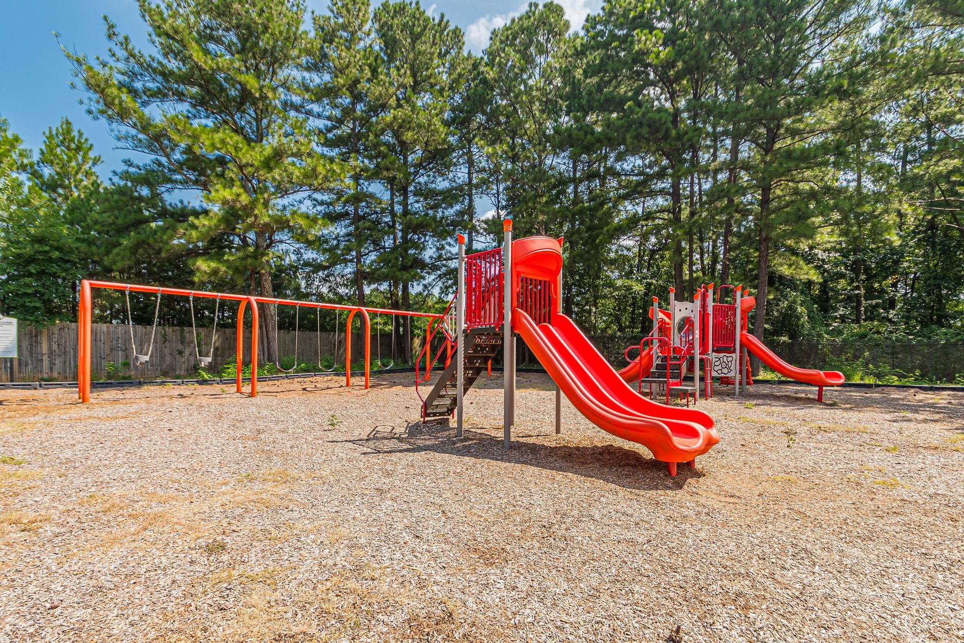 Empty playground with red slides and swings on a gravel lot, surrounded by trees.